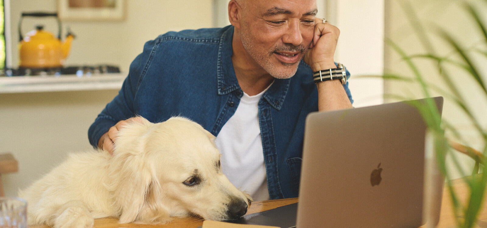 Middle-aged man smiling while using a laptop at home with a golden retriever beside him, featured in an article about overactive bladder in men, its symptoms, causes, and treatments.