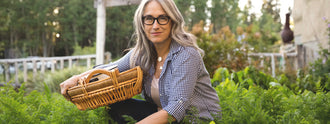 Woman with grey hair and glasses smiling in a garden while holding a wicker basket, featured in an article about what foods to eat or avoid on a menopause diet.
