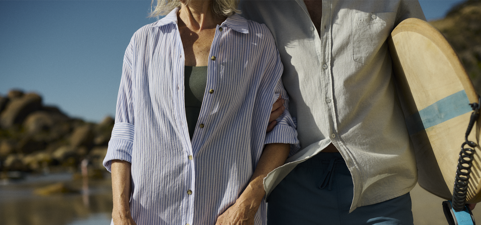 Close-up of an older couple walking arm-in-arm on the beach, one carrying a surfboard, featured in an article about essential tips for managing overactive bladder.