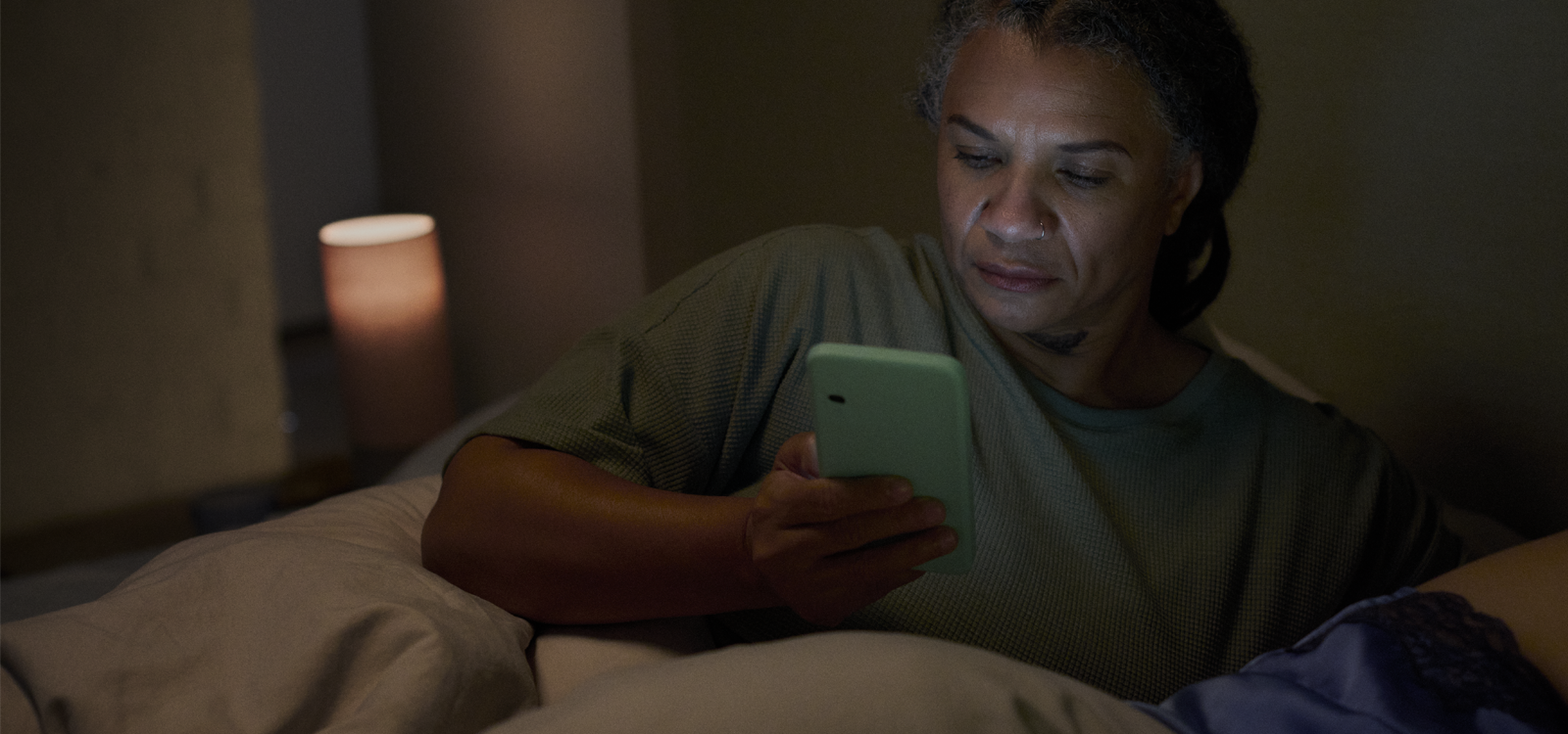 Middle-aged woman lying in bed at night, using a smartphone, with a soft bedside lamp in the background, illustrating how to manage incontinence while sleeping.